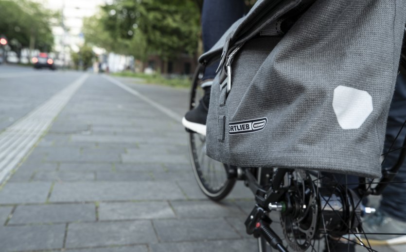 Un primer plano de una bolsa de bicicleta gris de Ortlieb sujeta a una bicicleta mientras la persona circula por un carril bici. Al fondo, se ve una carretera con coches y árboles.
