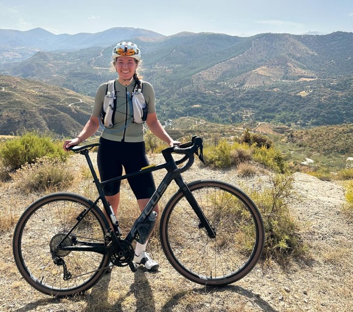 Eine junge Frau posiert mit ihrem Gravel-Bike auf einer Anhöhe vor einer bergigen spanischen Landschaft. Der Himmel ist wolkenlos