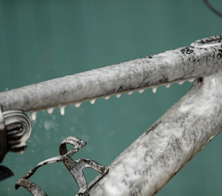 Nahaufnahme eines Fahrradrahmens, der mit Reinigungs­schaum bedeckt ist. Schaum tropft vom Oberrohr herab, während der Rahmen auf einem Montageständer fixiert ist.