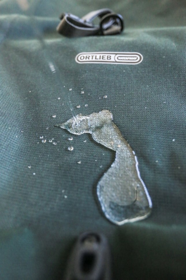 Nahaufnahme der grünen Oberfläche einer Ortlieb Back-Roller Urban Fahrradtasche mit Wassertropfen, die die wasserdichte Eigenschaft zeigen.