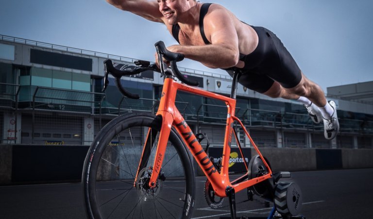 Ein Rennrad mit montiertem Indoor Trainer steht auf der Startposition einer Rennstrecke. Ein halbnackter Mann liegt bäuchlings auf dem Sattel in Superman-Pose.