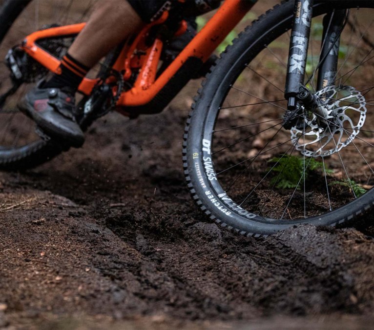 Rueda delantera de una bicicleta de montaña rodando por un sendero forestal con barro.