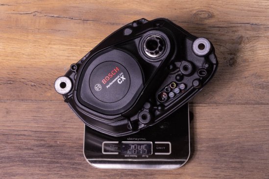 A Bosch CX e-bike drive on an electronic kitchen scale on a dark wooden floor. Display shows 2,845 grams