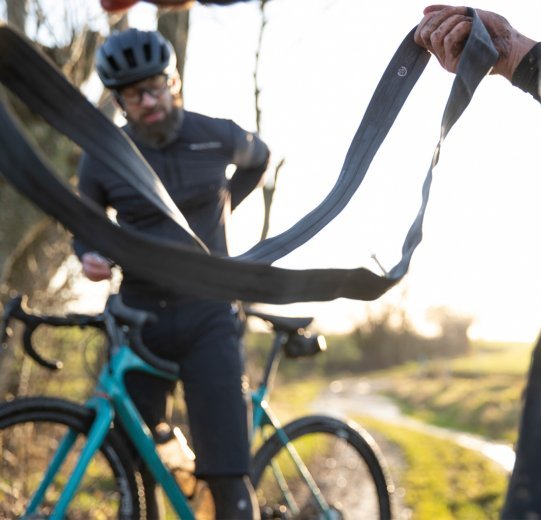 Two bikers deal with a puncture during a tour. While one pulls out a tube, the other searches in his back pocket for a CO2 cartridge.