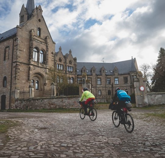 2 Teilnehmer fahren an einem alten Schloss vorbei über Kopfsteinpflaster.