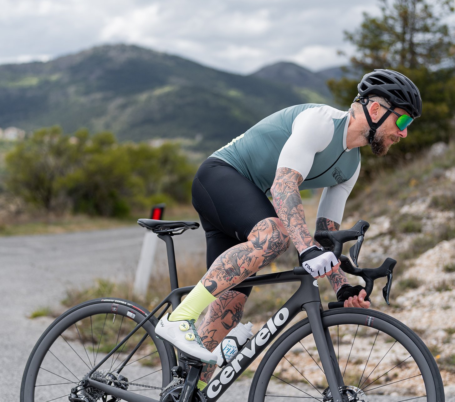 Mann mit Tattoos und Fahrradbekleidung fährt auf einem Cervélo-Rennrad eine kurvige Bergstraße hinauf. Er trägt einen Helm, Sonnenbrille, Radtrikot und neonfarbene Socken. Im Hintergrund sind bewaldete Hügel und ein bewölkter Himmel zu sehen