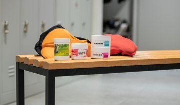 La photo montre 4 crèmes anti-irritations différente de Elite, Assos et Eule devant deux cuissards posés sur un banc dans les vestiaires pour les hommes chez bc. 