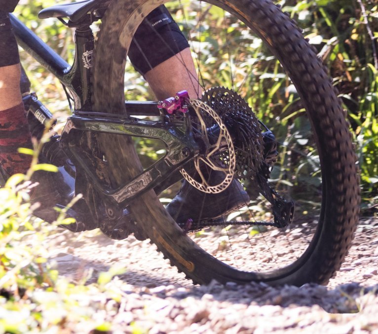 Gros plan d’un vététiste roulant sur un sentier forestier, avec la roue arrière, le disque de frein et le cadre bien visibles.