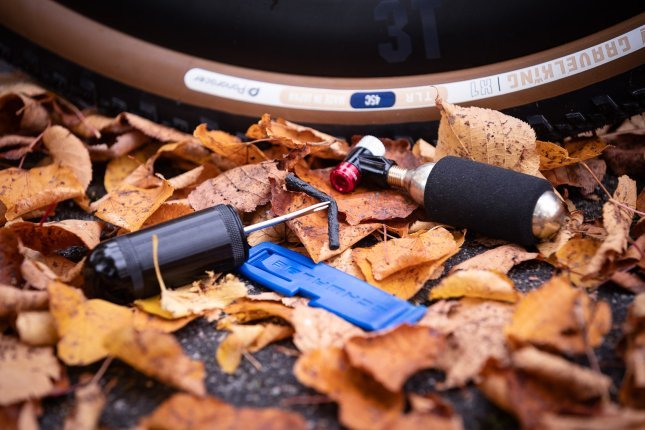 Eine CO2-Minipumpe, ein Reifenheber und ein Tubeless-Tool mit eingeklemmter Reifefensalami liegen vor einem Gravel-Reifen in buntem Herbstlaub 