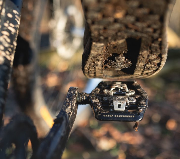 Detail photo: The sole of an MTB shoe, positioned just above a black OneUp Clip Pedal. In the blur, parts of a dark mountain bike and an autumn forest floor are visible.