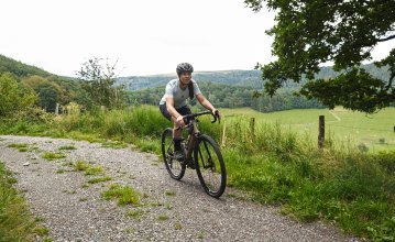 Susi aus dem Social Media-Team von bc fährt mit einem bc original Flint Gravelbike über eine Schotterpiste, umgeben von grüner, hügeliger Landschaft. 