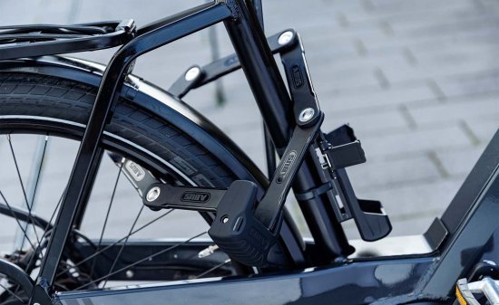 Candado plegable asegurando la rueda trasera y el cuadro de una bicicleta a un punto fijo; fijado de forma compacta al cuadro.