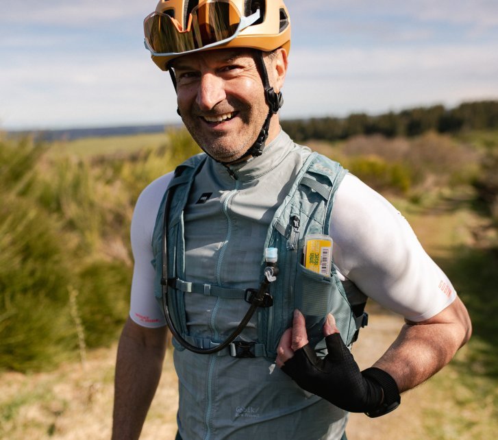 Ein ca. 50 Jahre alter Mann lacht in die Kamera.Er trägt einen Fahrradhelm, ein weißes Kurzarmtrikot und eine blaugraue Weste mit Taschen.