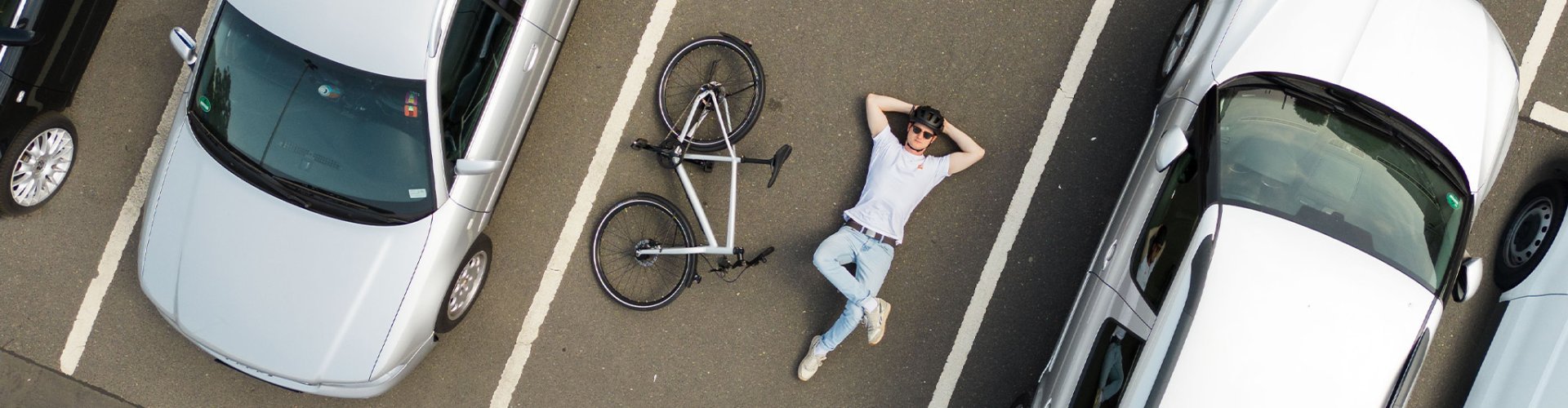 A person is lying relaxed on the ground of a parking lot next to a bicycle, which is also on the ground, between two parked cars.
