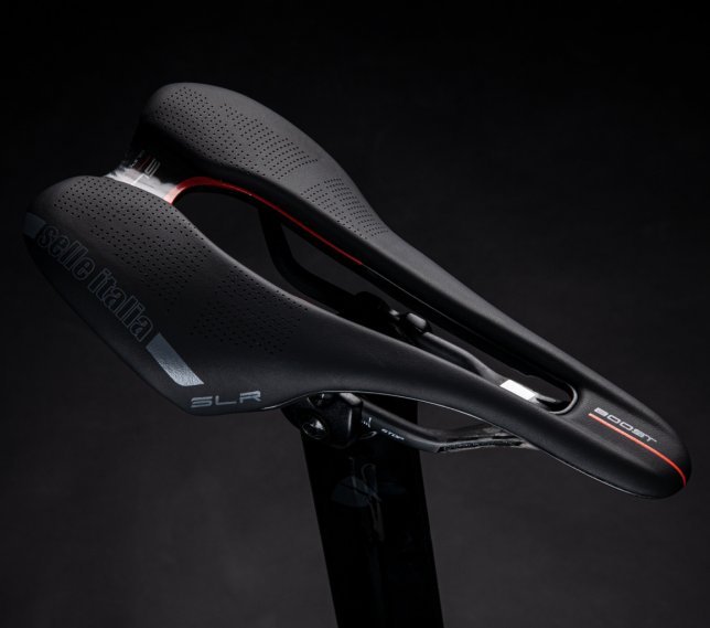 Close-up of a black Selle Italia SLR Boost bike saddle with red accents. The saddle features a perforated design and is shown against a dark background, with the "Selle Italia" and "SLR Boost" markings visible.