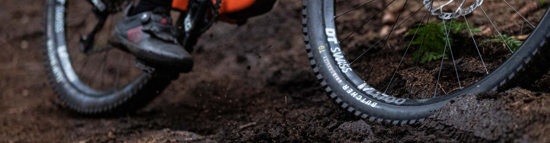 Rueda delantera de una bicicleta de montaña rodando por un sendero forestal con barro.