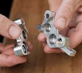 Dos manos de hombre sostienen las dos mitades de una pinza de freno Trickstuff plateada. En el interior se ven los cuatro taladros cilíndricos para los pistones de freno.