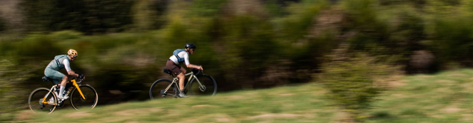 Deux cyclistes gravel roulent rapidement à travers une prairie vallonnée, arrière-plan flou soulignant le mouvement.