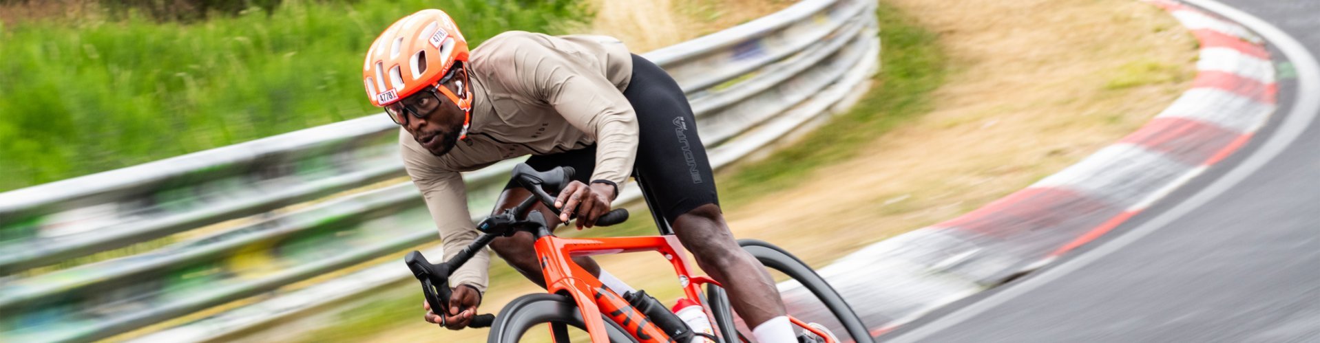 Road cyclist using drop handlebars leans low into a fast corner on a paved track, guardrail alongside and motion blur emphasizing speed and dynamic riding position.