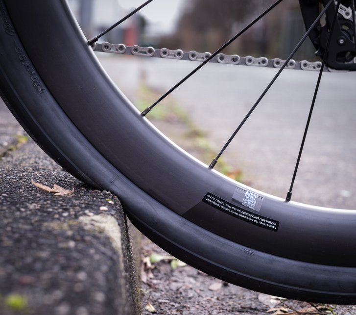 Detailfoto: Das Hinterrad eines Rennrades rollt einen Bordstein hoch. Man sieht, wie der Reifen von der Steinkante in Richtung Felge eingedrückt wird. 