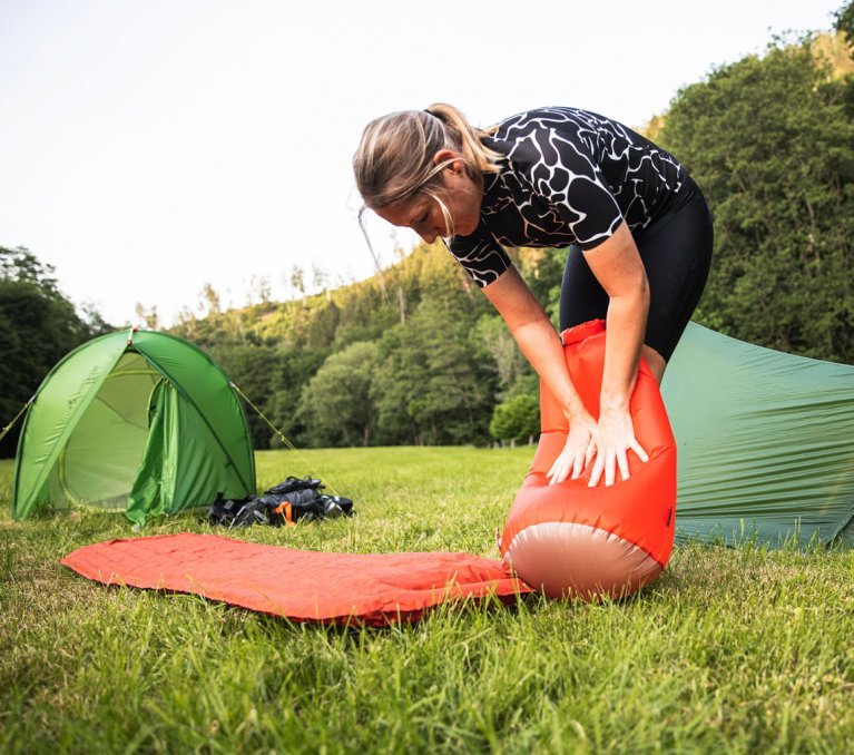 2 tentes VAUDE montées sur une prairie entourée d'arbres. Au premier plan, un matelas de camping est rempli d'air à l'aide d'un sac de gonflage.