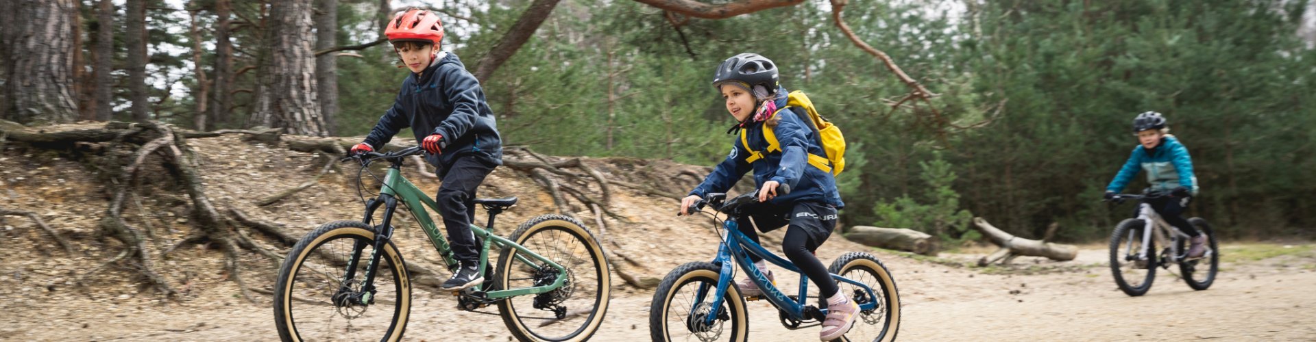 Three children ride on SUPURB and Specialized kids mountain bikes along a forest path.