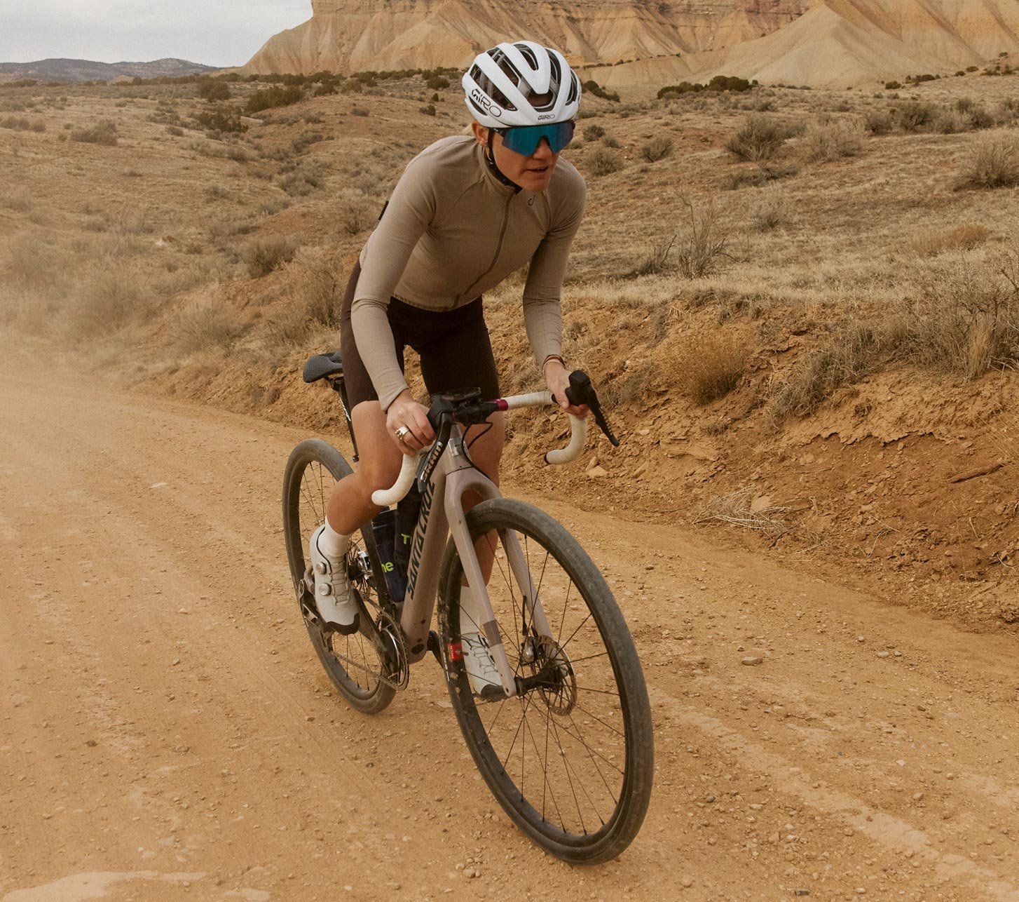 Gravel-Fahrerin mit weißem Giro Fahrradhelm fährt auf staubiger Schotterstraße durch eine weite Wüstenlandschaft.