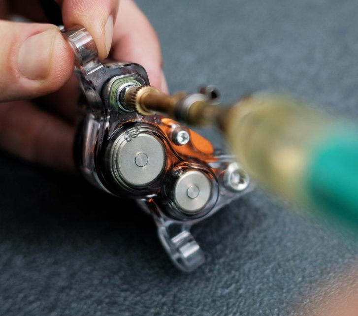 Una pinza de freno de 4 pistones de Trickstuff con carcasa transparente se está llenando con una jeringa. Se pueden ver claramente los pistones de acero y dos burbujas de aire.