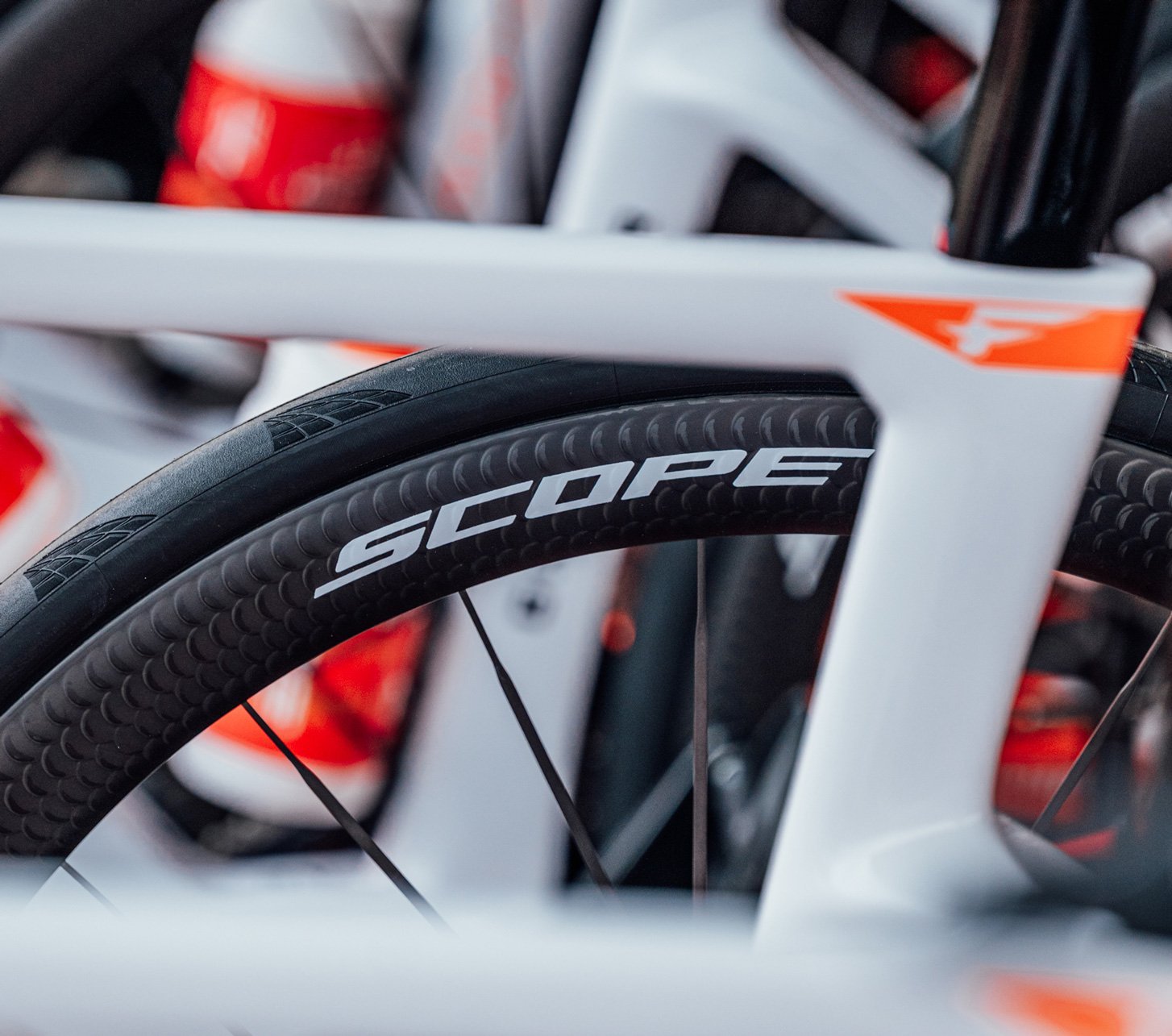 Close-up of a Scope Cycling carbon wheel on an INEOS Grenadiers road bike with a white and red frame and black aero rim.