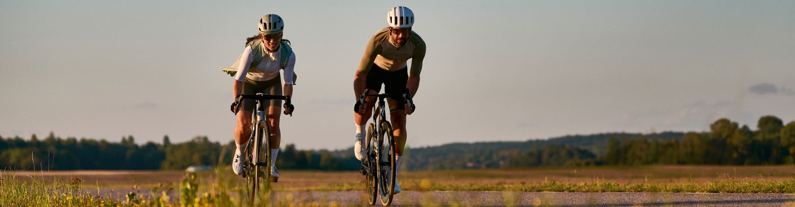Deux cyclistes sur route portent des vêtements de cyclisme Craft et roulent côte à côte sur une route de campagne dans un paysage ouvert au coucher du soleil.