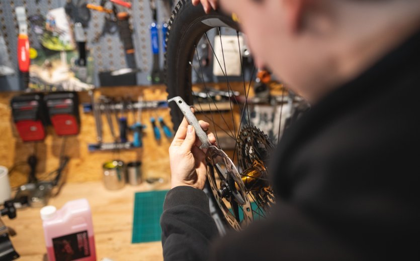 Blick über die Schulter eines Mechanikers: Seine Hand hält ein Bremsscheiben-Richtwerkzeug an die Bremsscheibe eines MTB-Laufrads.
