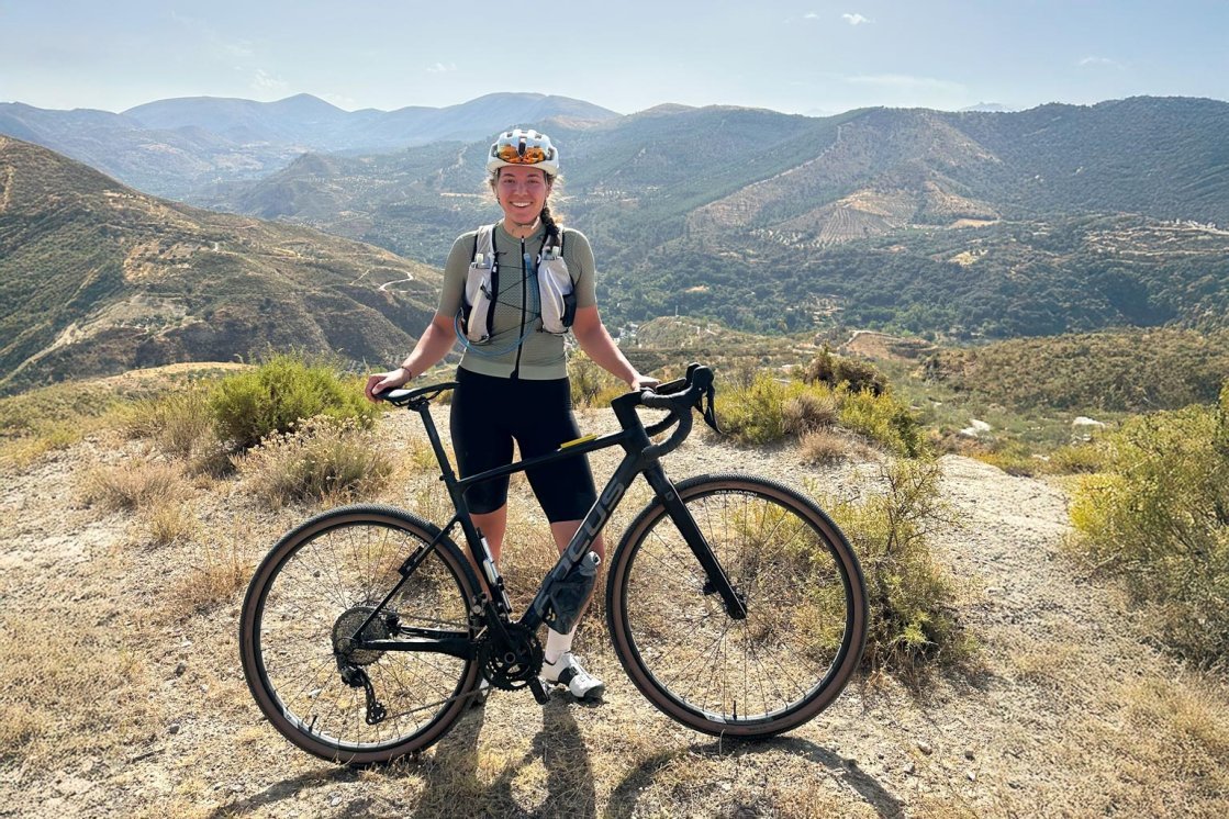 Eine junge Frau posiert mit ihrem Gravel-Bike auf einer Anhöhe vor einer bergigen spanischen Landschaft. Der Himmel ist wolkenlos