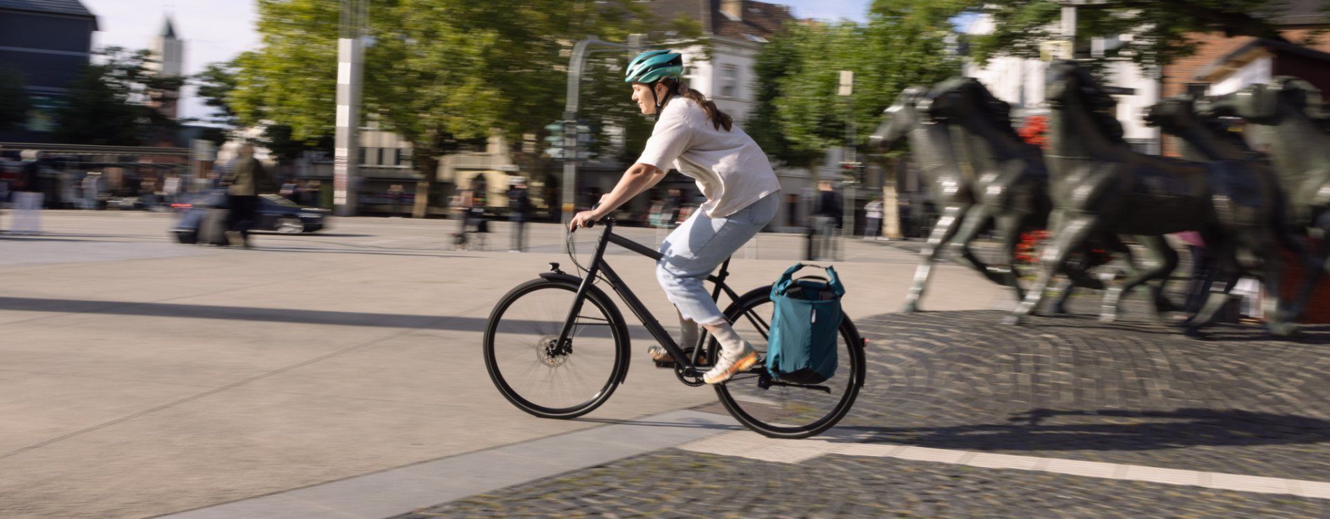 A person wearing a helmet rides a bicycle across an urban square; in the background, pedestrians and several horse sculptures can be seen.