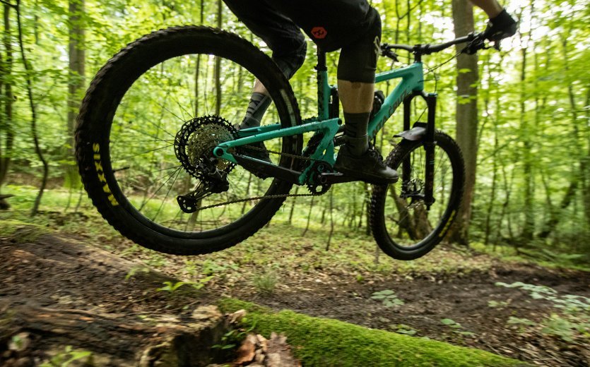 A mountain biker rides his Santa Cruz over a trail in a forest. He takes off. 