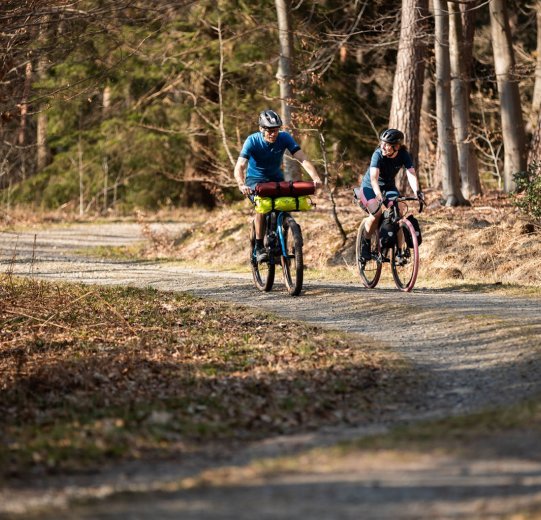 Rainer und Svenja von bc fahren mit ihren bepackten Bikes über einen Waldweg. Es ist sonnig.