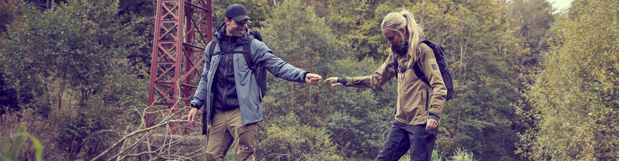 Two hikers wearing outdoor clothing and backpacks cross a stream beneath an iron bridge