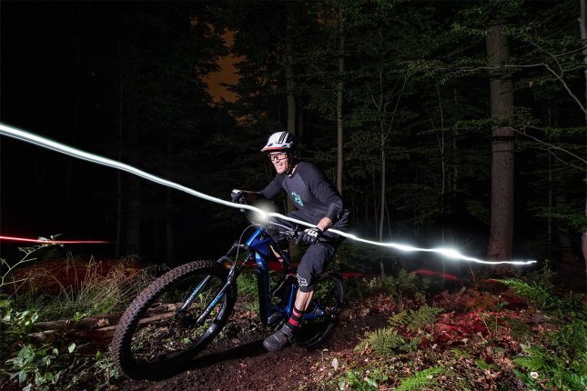 Un ciclista de E-mountain bike pedalea de noche en un sendero del bosque, iluminado por una luz en el casco, con trazos de luz marcando el camino y árboles rodeando el entorno.