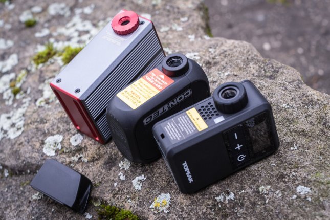 Drei kleine Akku-Pumpen liegen nebeneinander auf einem mit Flechten bewachsenen Stein. Darunter liegt ein SRAM AXS-Akku zum Größenvergleich