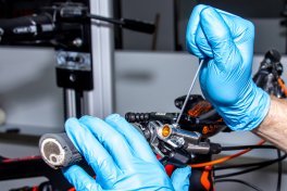 A hand wearing blue gloves unscrews the bleed screw on a bicycle brake lever while the bike is mounted on a stand.