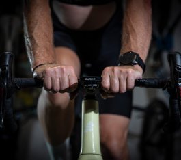 Dos manos de hombre agarran el manillar de una bicicleta de ruta. El ambiente parece oscuro, iluminado por un flash, como en un sótano.