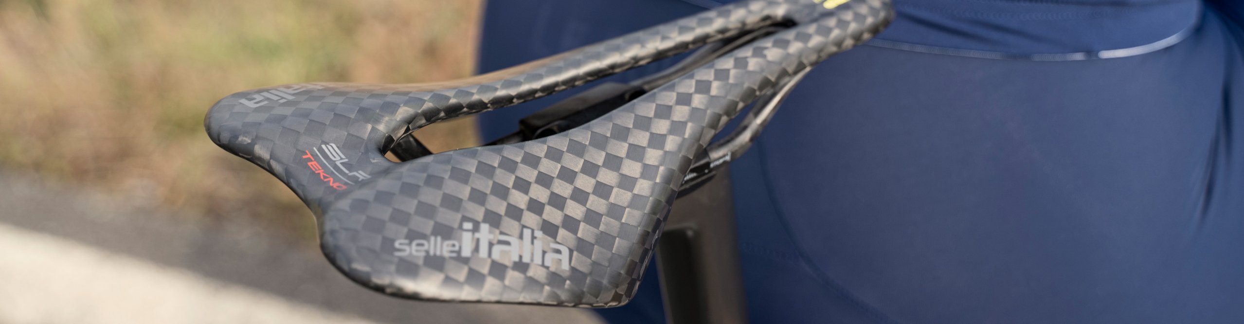Gros plan sur une selle carbone Selle Italia montée sur un vélo de route, à côté d’une personne cycliste en cuissard bleu.