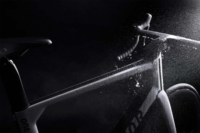 Nahaufnahme eines Fahrradrahmens im Dunkeln, auf dem Wasser abperlt. Tropfen laufen vom Oberrohr und Steuerrohr ab. Die Oberfläche wirkt glatt und hydrophob, während feiner Sprühnebel im Licht sichtbar ist.
