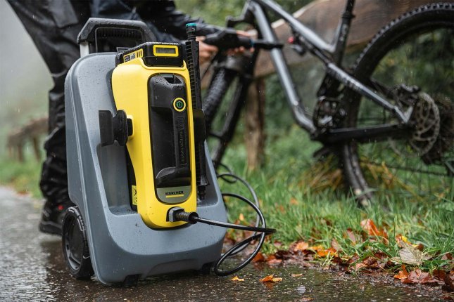 Mobiler Mittelruckreiniger von Kärcher steht auf nassem Asphalt neben einem Mountainbike. Eine Person reinigt das Fahrrad im Freien, Laub liegt am Boden. Das kompakte Reinigungsgerät wird für eine schonende Fahrradwäsche eingesetzt.