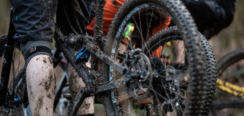 A very dirty MTB shorts and a very dirty man's calf next to a very dirty mountain bike.