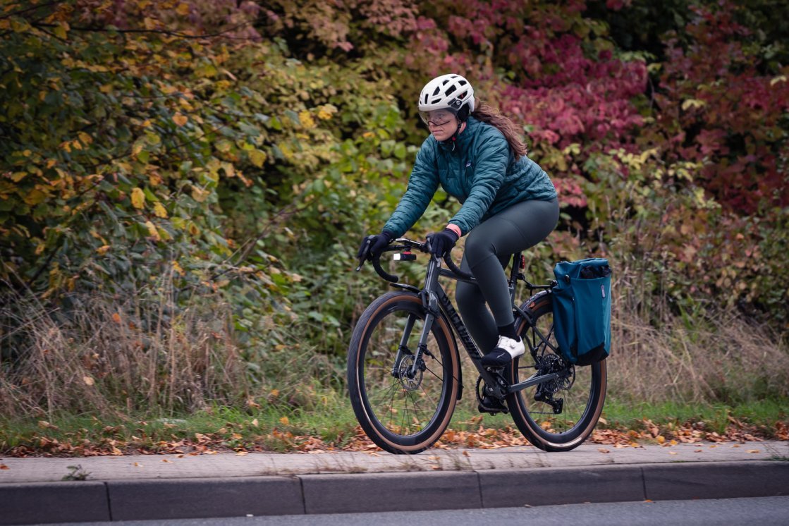 A young woman in sea-green winter clothing rides a graphite-grey gravel bike through an autumnal landscape.