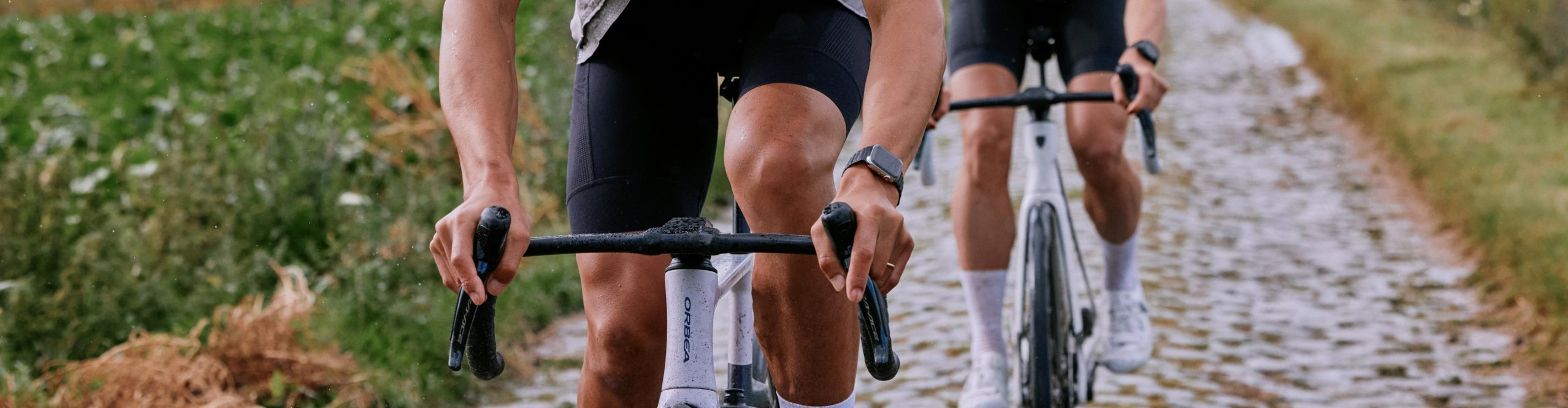 Zwei Rennradfahrer fahren bei nassem Wetter auf Rennrädern mit PRO Lenkern über einen Kopfsteinpflasterweg.