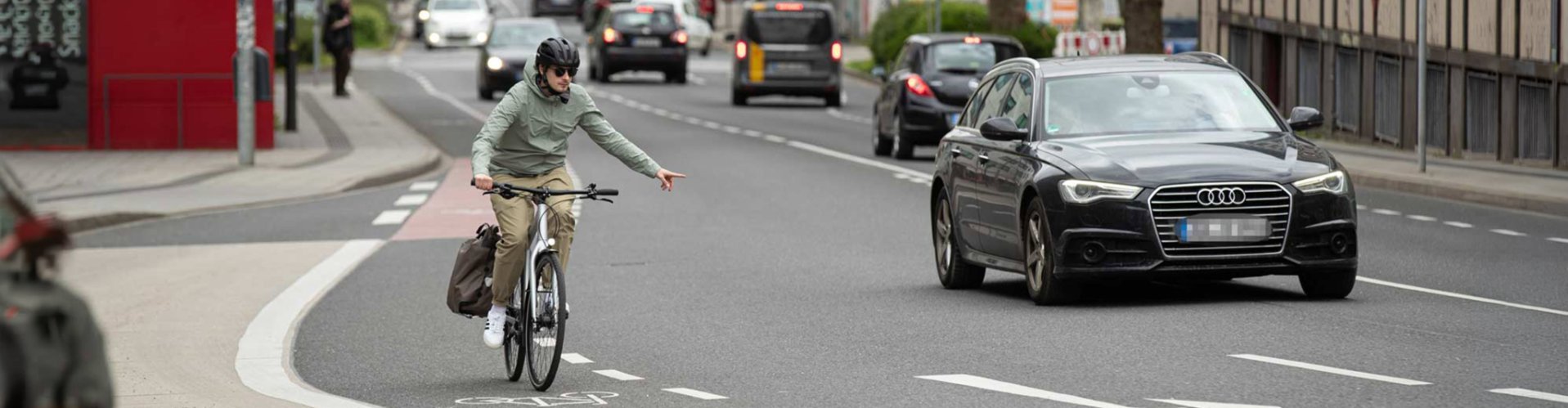Un cycliste portant un casque roule sur une piste cyclable balisée dans la circulation urbaine et signale d'un geste de la main qu'il change de voie, alors que des voitures circulent sur la route.