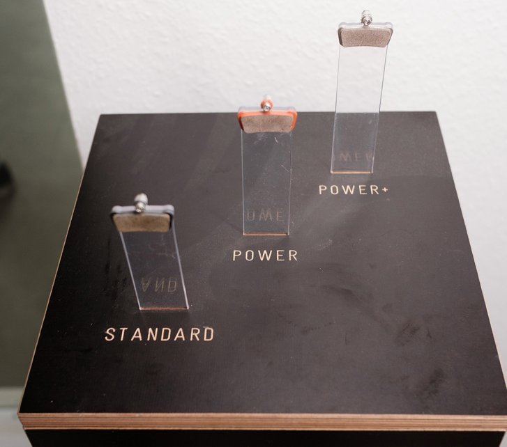 Three transparent tubes containing brake pads are placed on a black surface labeled with "POWER" and "STANDARD."