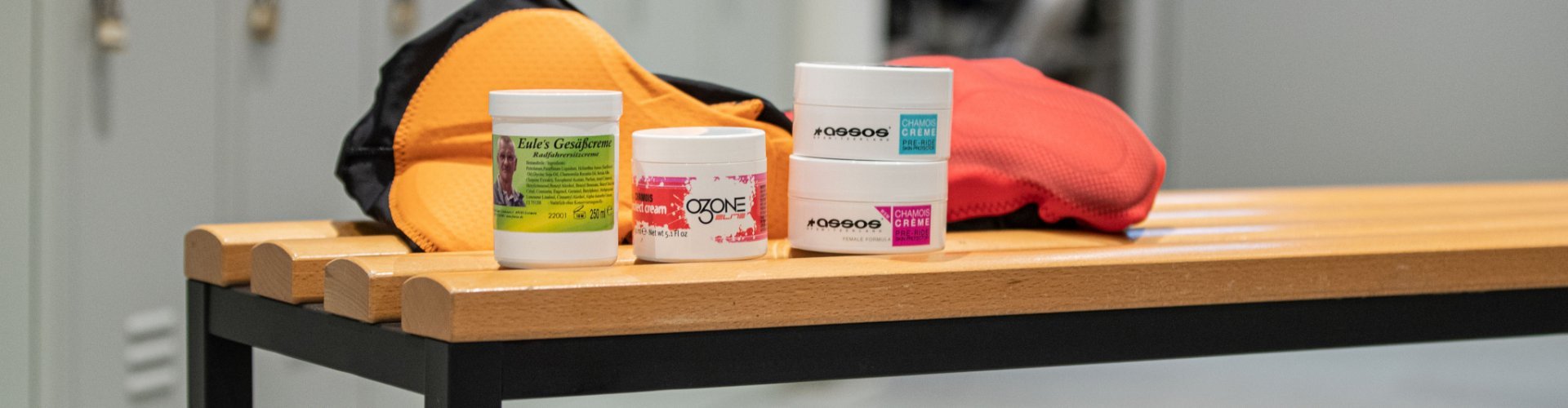 Pictured are 4 varieties of chamois cream from Elite, Assos and Eule in front of 2 bib shorts on a bench in the bc men's locker room.