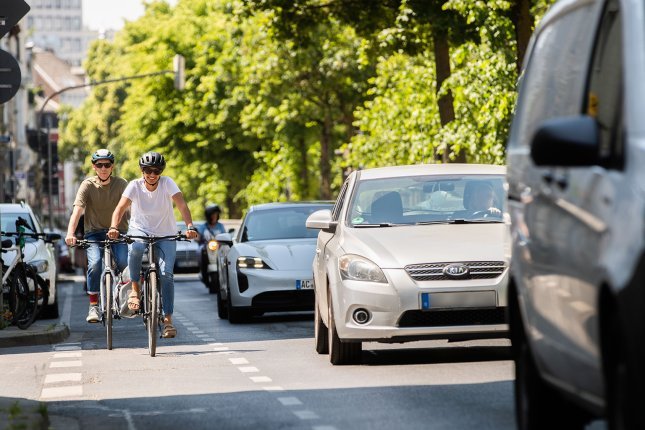 Two people are riding bicycles on a bike lane in a city street, while cars are stuck in traffic next to them. Trees and buildings are visible in the background.